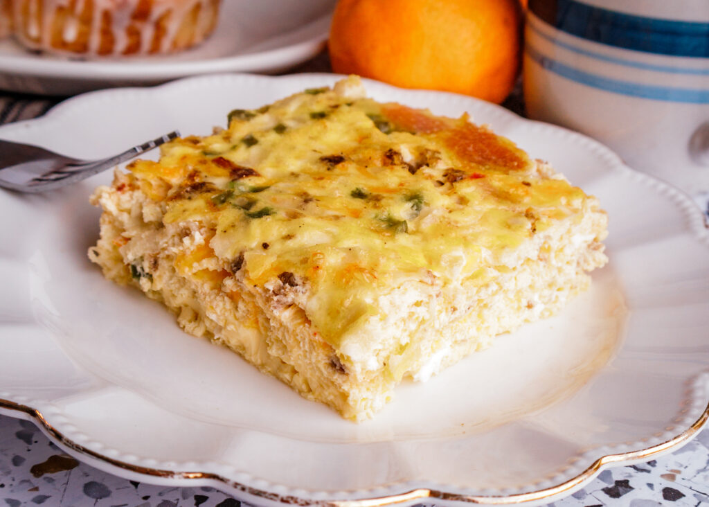 A gluten free serving of egg and sausage breakfast casserole on a white plate, served with fresh orange slices, a cinnamon roll, and a cup of coffee