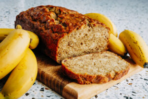 Slices of moist honey sweetened banana bread on a wooden board next to whole ripe bananas, made with whole cell crushed wheat and no refined sugar.