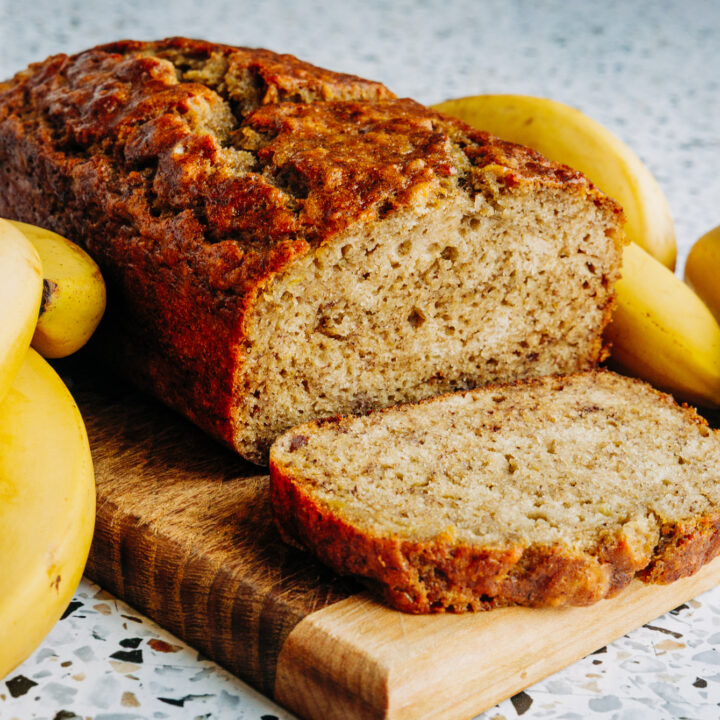 Slices of moist honey sweetened banana bread on a wooden board next to whole ripe bananas, made with whole cell crushed wheat and no refined sugar.