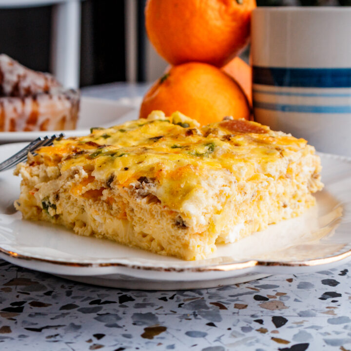 A square serving of egg and sausage breakfast casserole on a white plate, served with fresh orange slices, a cinnamon roll, and a cup of coffee