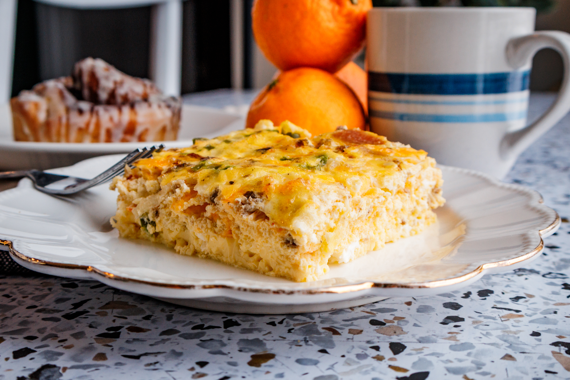 A square serving of egg and sausage breakfast casserole on a white plate, served with fresh orange slices, a cinnamon roll, and a cup of coffee