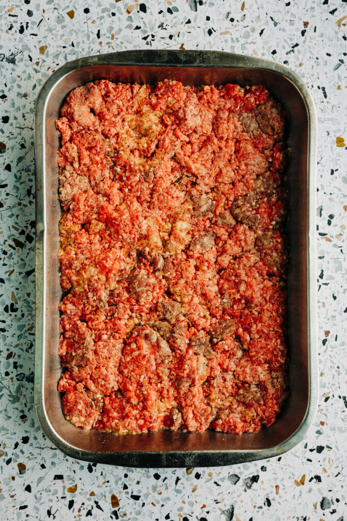 Raw ground beef seasoned and pressed evenly into a 9x13 metal baking pan for slider patties.